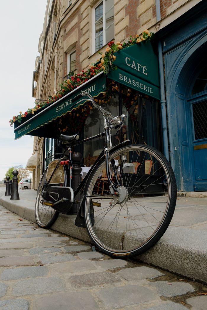 product-04 A classic bicycle parked beside a charming Parisian café and brasserie on a cobblestone sidewalk, showcasing urban charm.